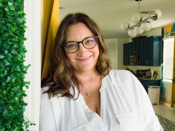 Cari Kaufman, Storyteller Wordsmith, specializing in Copywriting, from Lowell, Arkansas, head-and-shoulders portrait of a smiling woman with shoulder-length wavy brown hair and black-framed glasses, wearing a white blouse and small pendant necklace, leaning against a doorway beside a vertical green plant wall in a bright home kitchen.