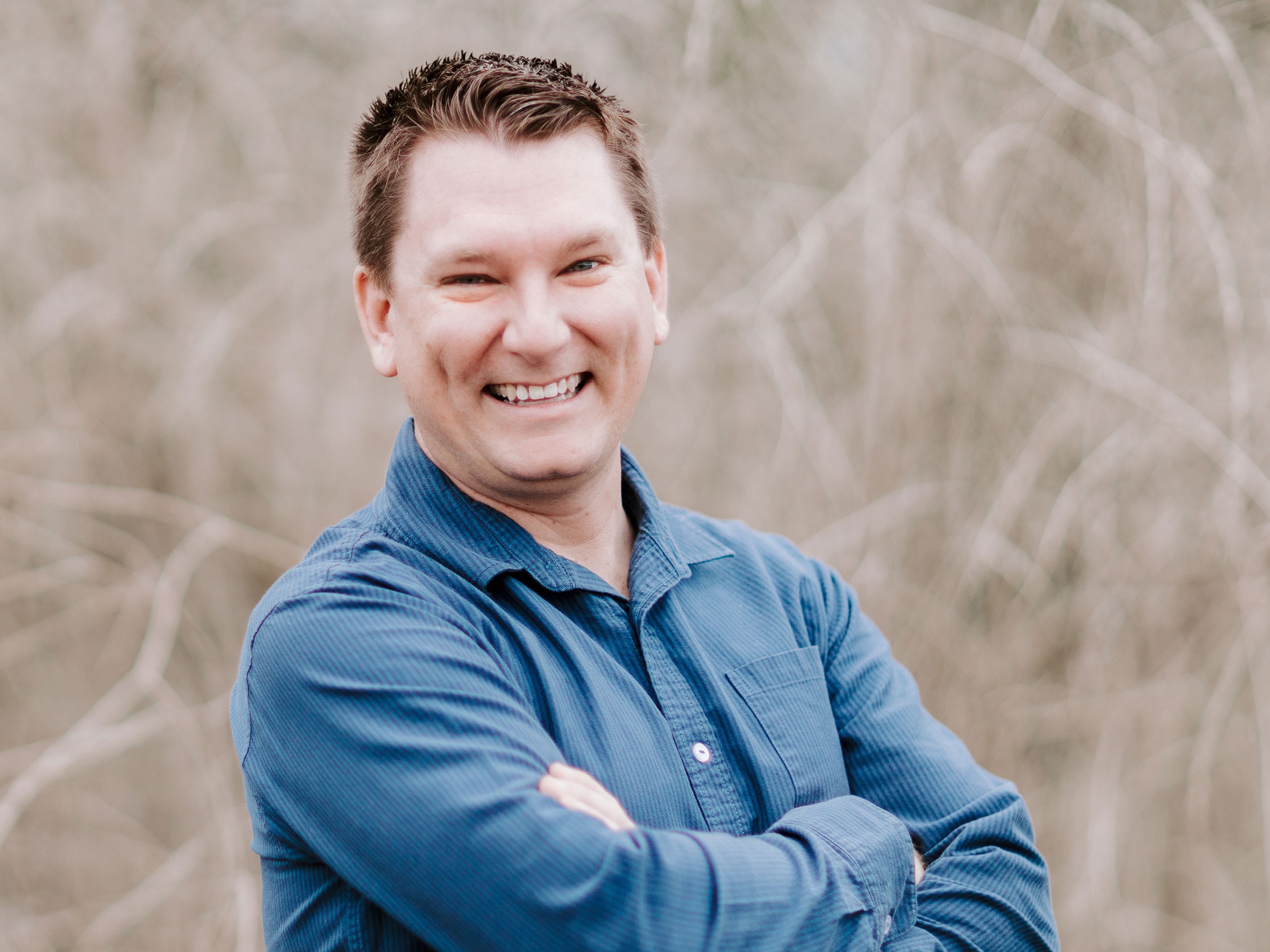 David Hepburn, Hepburn Creative, specializing in Marketing Strategy & Consulting, from San Diego, California, head-and-shoulders portrait of a smiling man with short brown hair wearing a blue button-up shirt with arms crossed against a softly blurred neutral outdoor background.