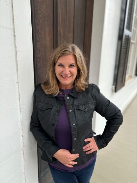 Stacey Marler, specializing in Marketing Content Strategist , from Roswell, Georgia, smiling in a waist-up standing portrait with shoulder-length light brown hair with subtle waves, wearing a black jacket over a purple top and dark jeans, leaning against a white column and dark wooden door with one hand on her hip.