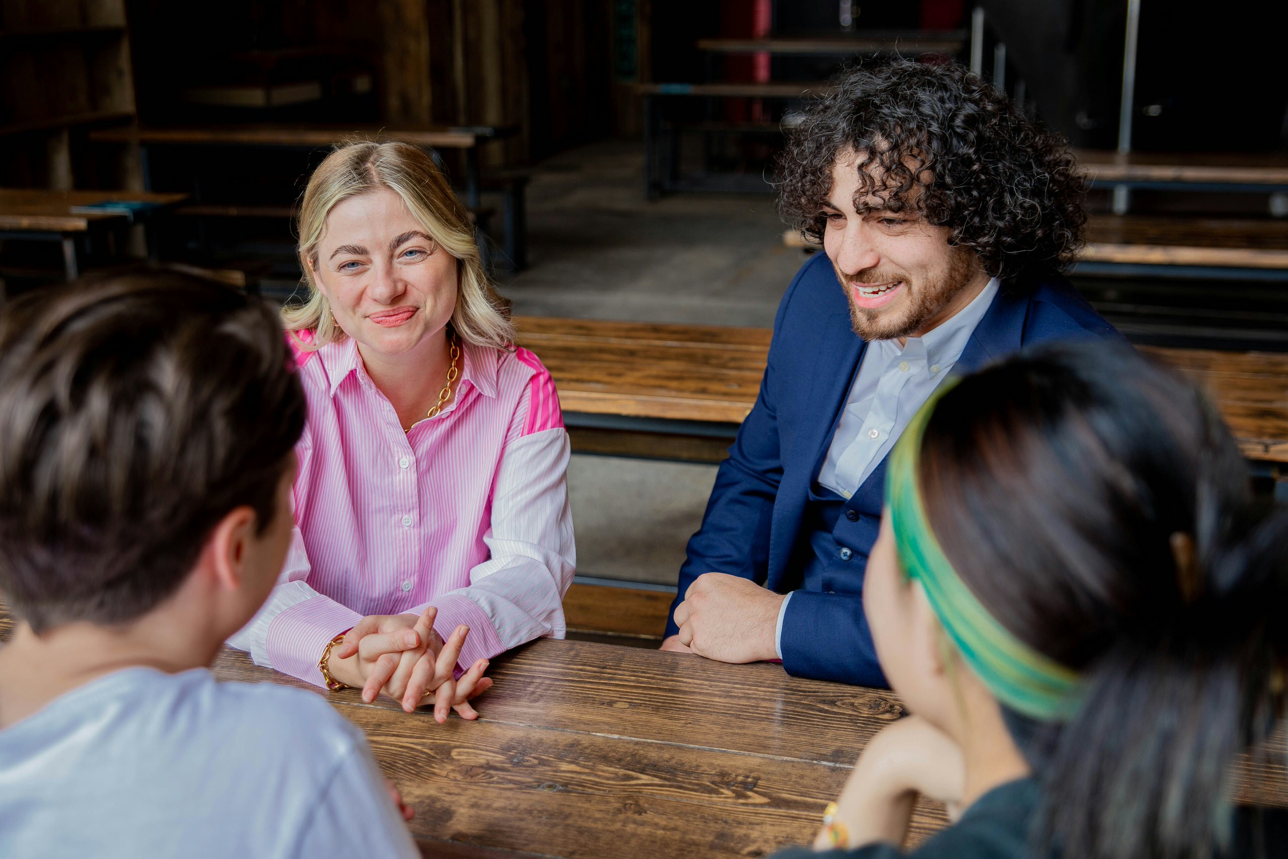 A woman with shoulder-length blonde hair in a pink and white blouse and a man with curly dark hair, a beard, and a blue suit are smiling and seated at a wooden table in a casual indoor setting with two other people.