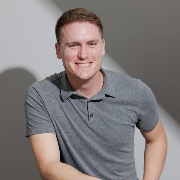 Jordan Ryskamp, Clarity Messaging, specializing in Web Strategy, from Virginia Beach, Virginia, head-and-shoulders portrait of a smiling man with short light brown hair and fair complexion wearing a gray polo shirt, seated and leaning slightly forward against a neutral gray wall with a diagonal band of light.
