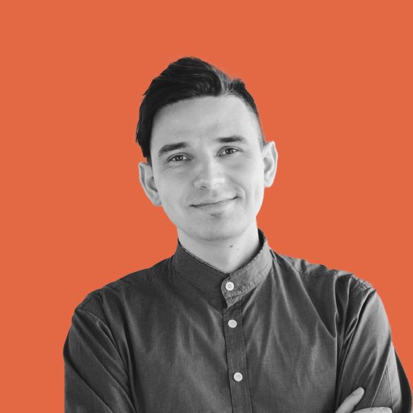 Māris Prindulis, StoryClicks, specializing in Graphic design, WordPress, StoryBrand, Riga, Latvia — head-and-shoulders portrait of a man with dark swept-back hair wearing a dark button-up band-collar shirt, smiling with arms crossed against a solid orange background.