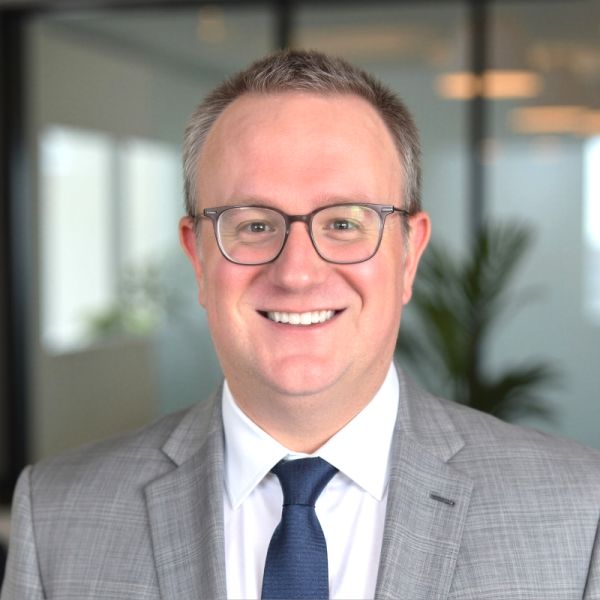 Jake Miller, specializing in Sales & GTM, from New Hampshire, head-and-shoulders professional portrait of a man with short graying hair and rectangular glasses, smiling, wearing a gray checked suit jacket, white dress shirt and navy tie, against a bright office background with a blurred glass wall and a plant.