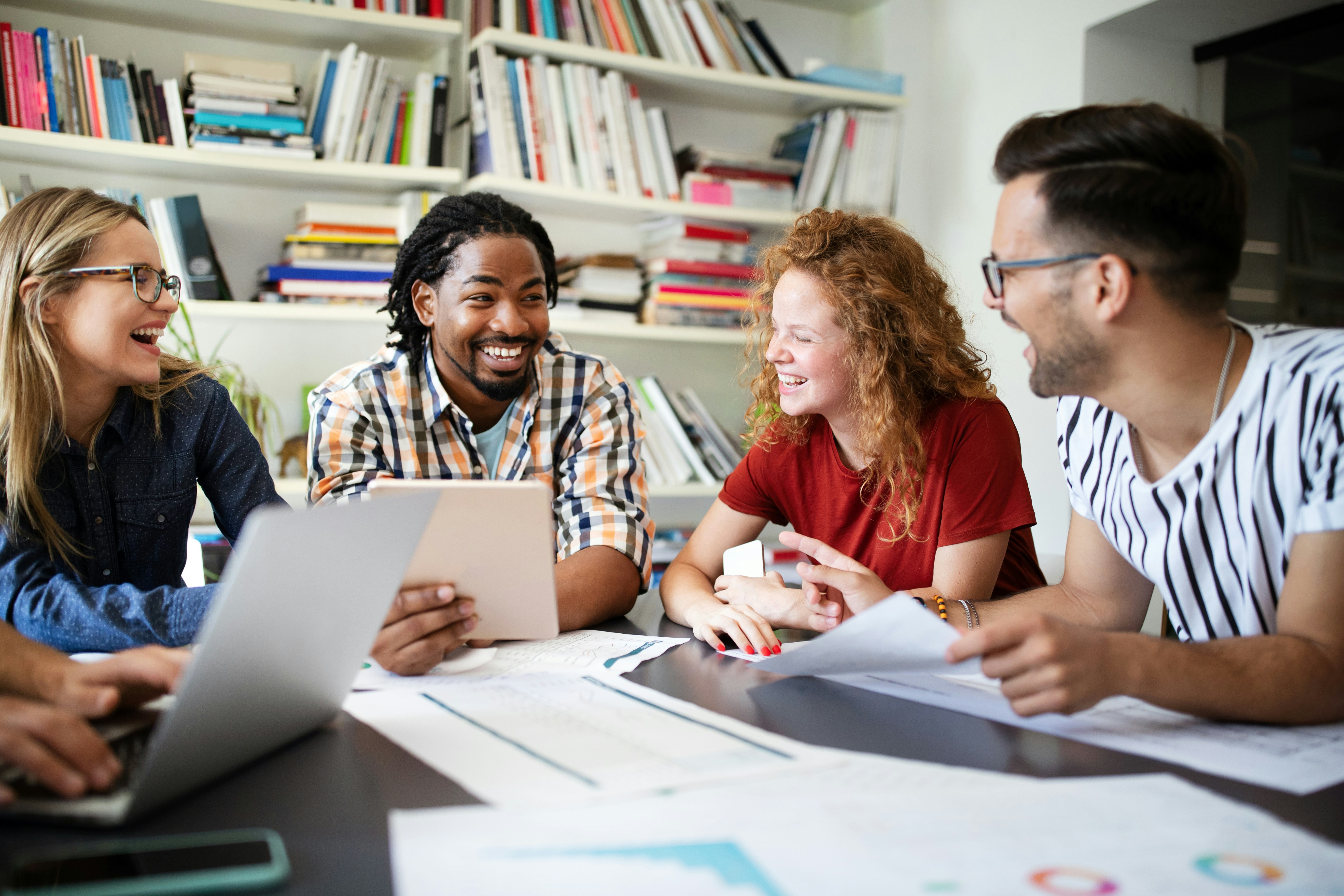 Vier junge Menschen arbeiten zusammen an einem Tisch in der Bibliothek und lachen dabei herzlich