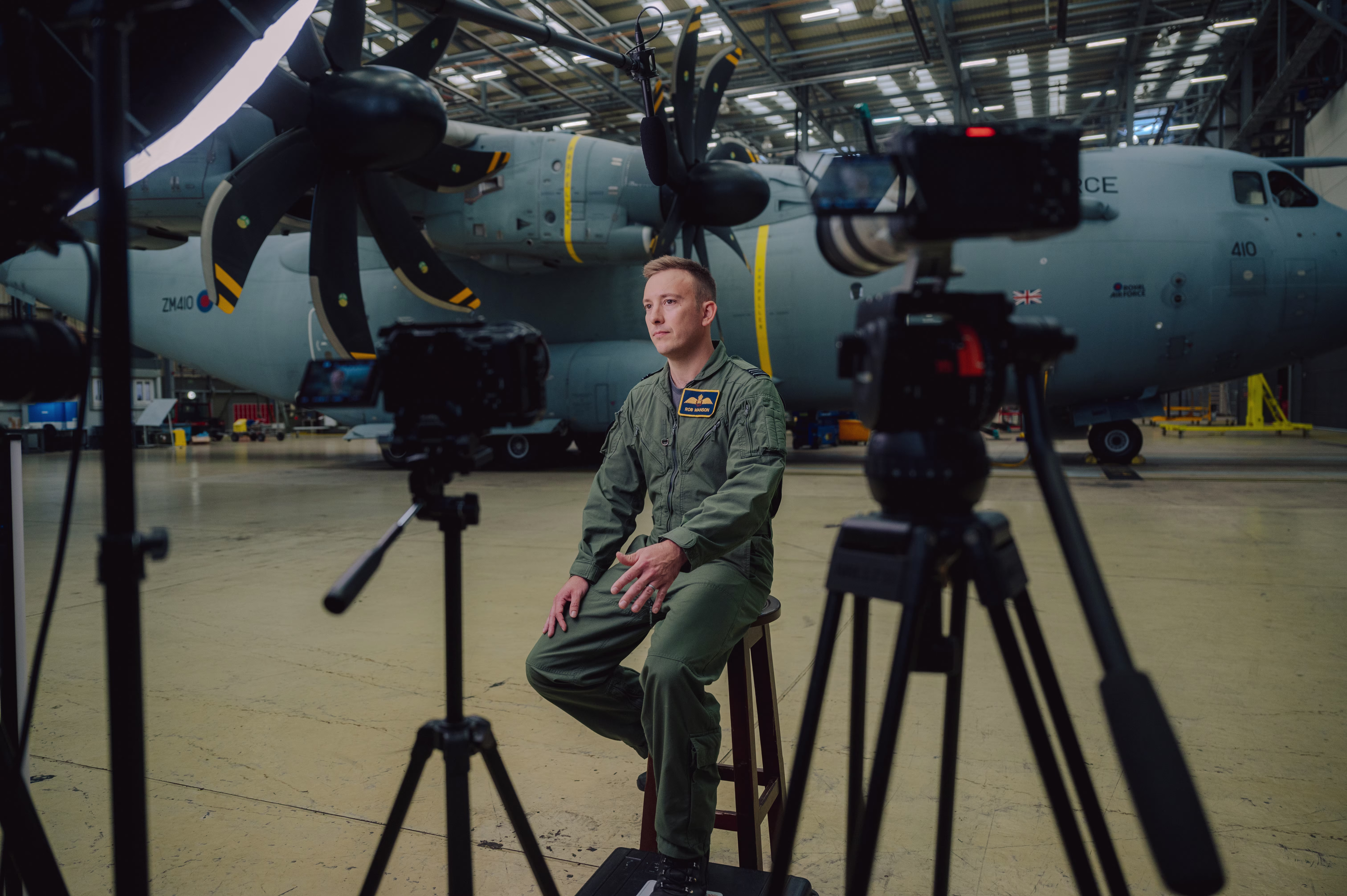 Sam Eckholm in green flight suit sitting on a stool in front of a large military aircraft during a video recording inside a hangar.