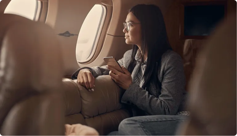 Woman with glasses sitting inside a private jet, holding a smartphone and looking out the window.