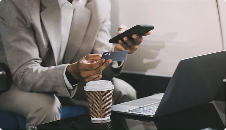 Person in a gray suit holding a credit card and using a smartphone next to an open laptop and a coffee cup on a table.