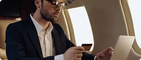 Man in glasses and suit using a laptop while holding a credit card inside an airplane.