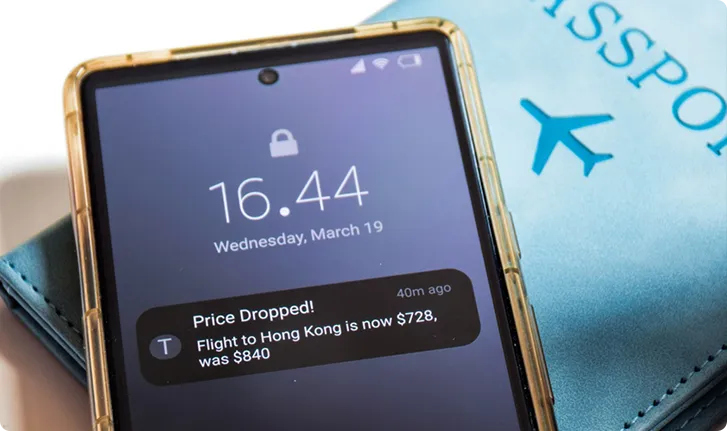 Smartphone screen showing a notification about a dropped flight price to Hong Kong from $840 to $728, next to a blue passport.