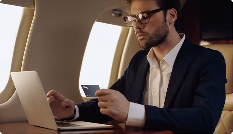 Person in a gray suit holding a credit card and using a smartphone next to an open laptop and a coffee cup on a table.