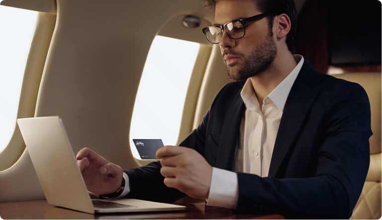 Person in a gray suit holding a credit card and using a smartphone next to an open laptop and a coffee cup on a table.