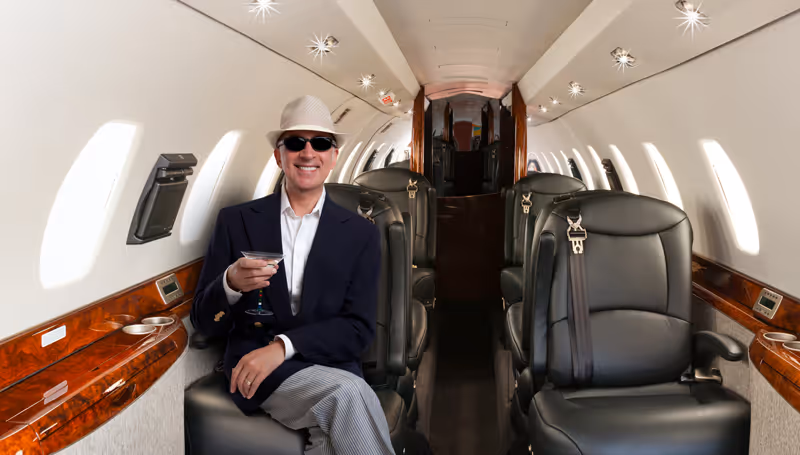 Man in sunglasses and hat sitting with a drink inside a luxury private jet cabin with leather seats and wood paneling.
