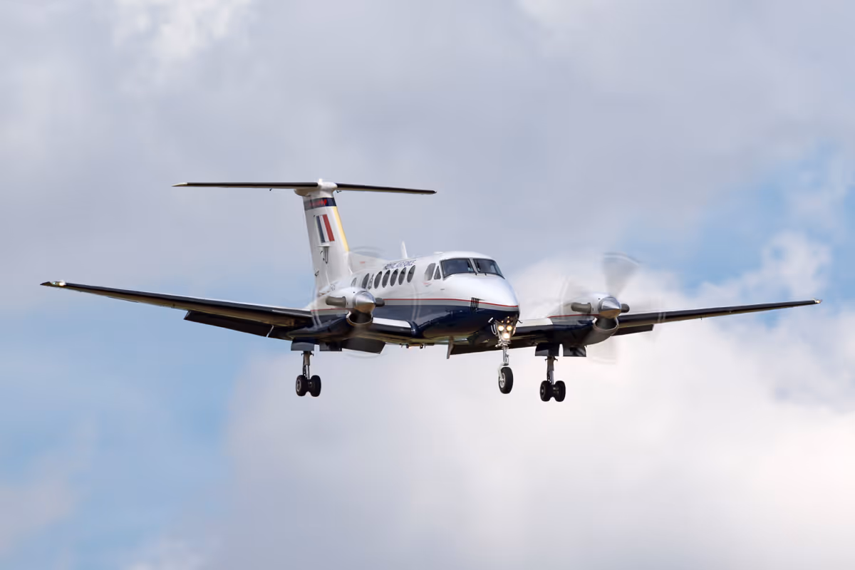 White twin-engine propeller aircraft approaching to land with landing gear extended against a cloudy sky.