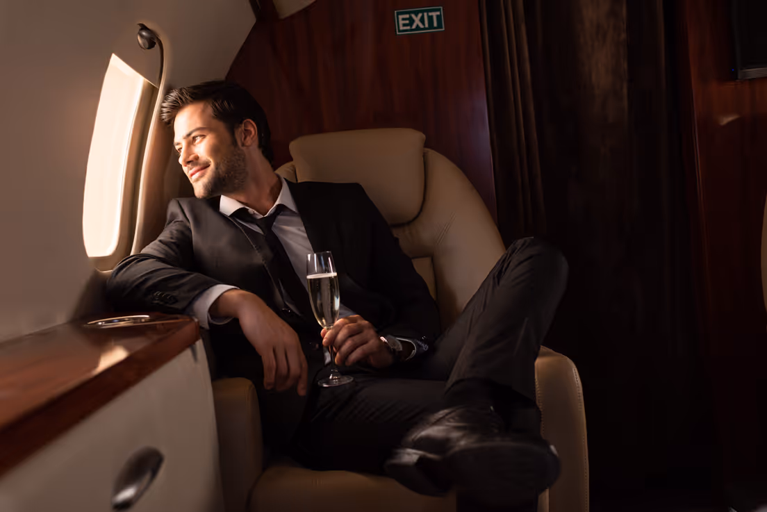 Man in a black suit sitting in a luxurious airplane seat, holding a glass of champagne and looking out the window with a smile.