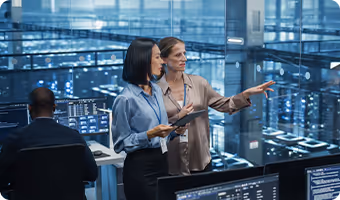 Two professional women in a modern data center, one holding a tablet and the other pointing while discussing, surrounded by server racks and monitors.