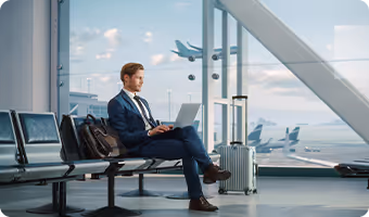 Businessman in a suit sitting in an airport lounge using a laptop with suitcases nearby and airplanes visible through large windows.