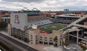 Aerial view of a large football stadium with University of Arizona logo on the side, surrounded by city buildings under a cloudy sky.