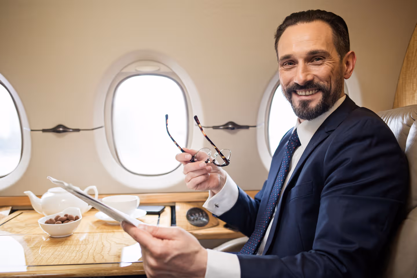 Smiling man in a suit holding glasses and a newspaper, seated inside a private jet with windows and a tea set on the table beside him.