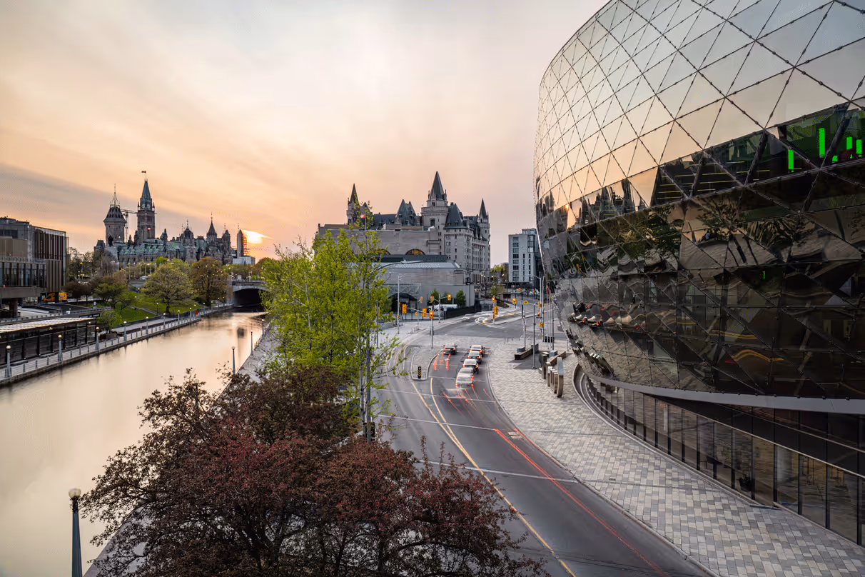 Cityscape at sunset with a river on the left, historic castle-like buildings in the distance, green and red trees, and a modern glass building on the right reflecting the surroundings.