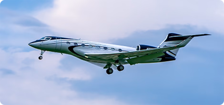 Silver and black private jet flying with landing gear extended against a cloudy sky.