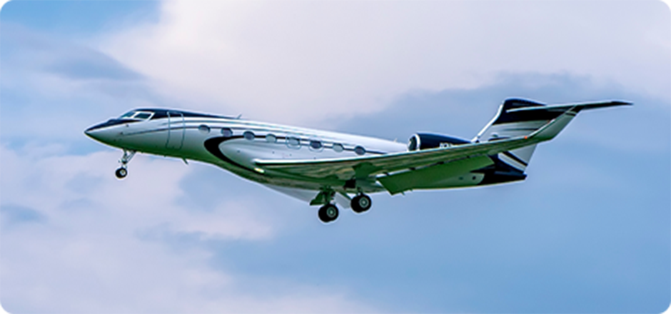 Silver and black private jet flying with landing gear extended against a cloudy sky.