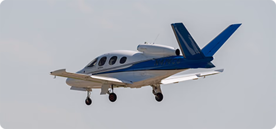Small white and blue private jet airplane flying with landing gear extended against a pale sky.