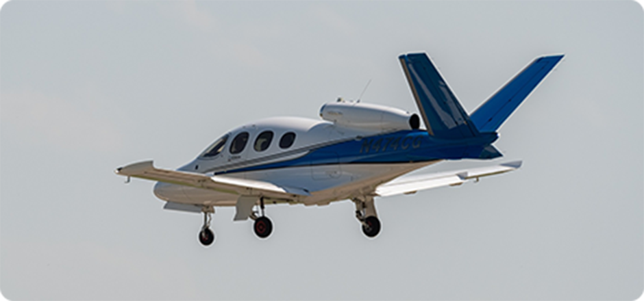 Small white and blue private jet airplane flying with landing gear extended against a pale sky.