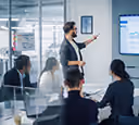 A man giving a presentation to colleagues in a modern glass-walled office meeting room.