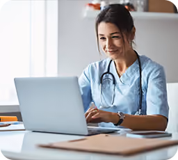 Smiling female doctor wearing scrubs and a stethoscope working on a laptop at a desk in a bright office.