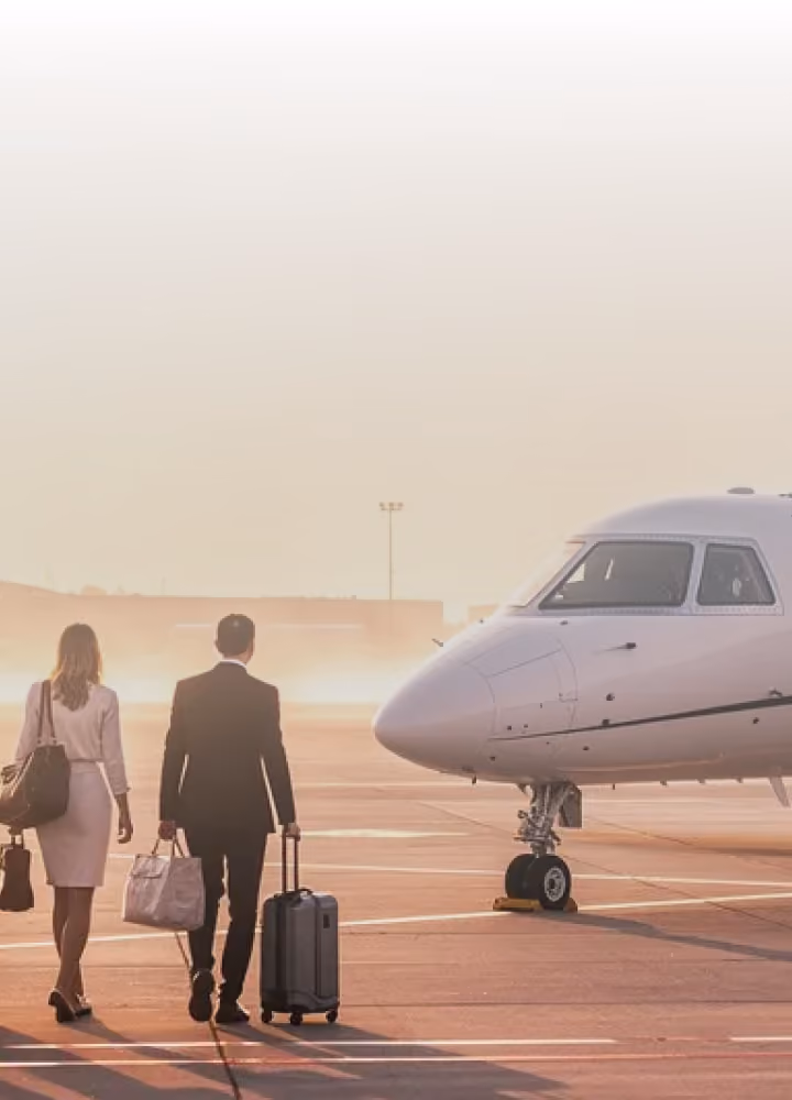 Businessman and businesswoman walking toward a private jet on the runway with luggage at sunrise.