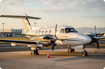 Small white twin-engine propeller airplane parked on airport tarmac near runway at sunset.
