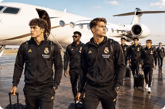 Real Madrid players walking on tarmac near a private jet wearing black tracksuits and carrying luggage.