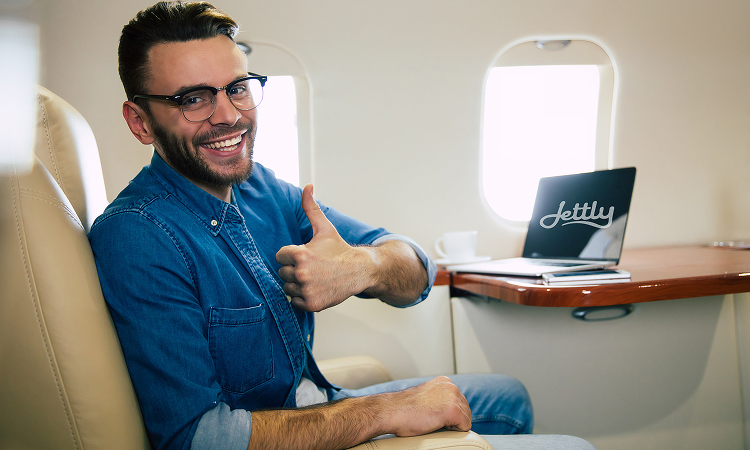 Smiling man in glasses giving thumbs up while seated on a private jet with a laptop on the table.
