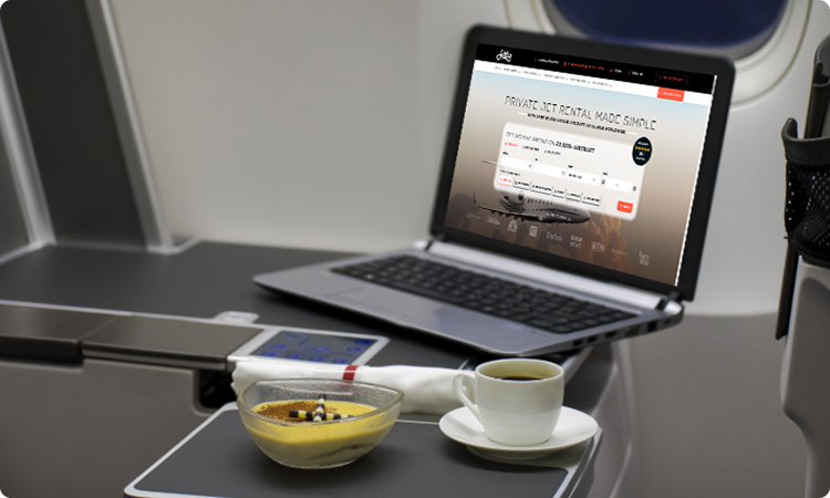 Laptop on airplane tray table displaying private jet rental site with coffee and dessert nearby.