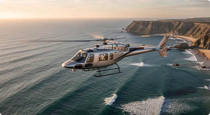 Silver helicopter flying over ocean waves near a rocky coastline at sunset.