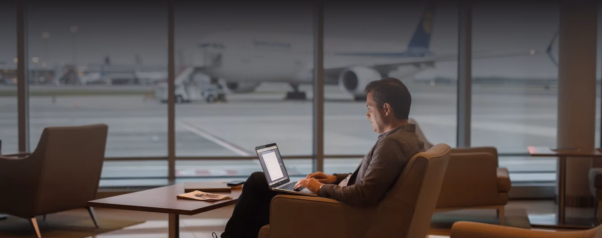 Man sitting in airport lounge chair working on a laptop with airplane visible outside window.