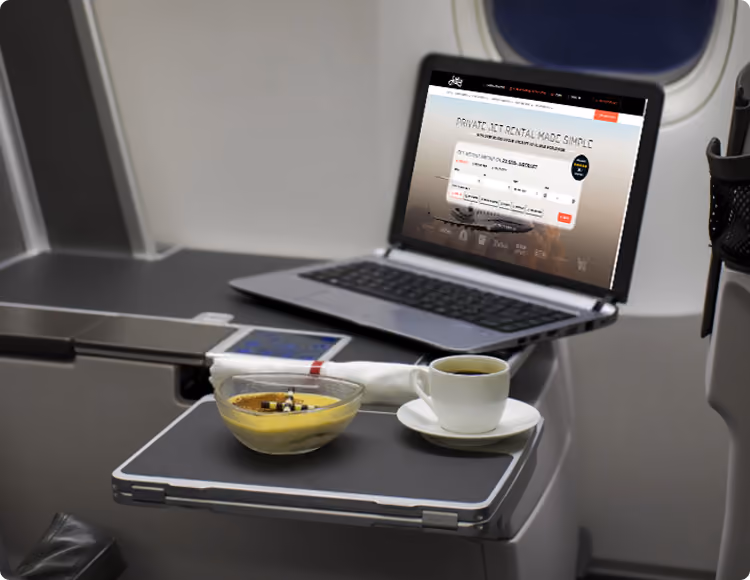 Tray table on airplane with coffee cup, dessert bowl, napkin, and open laptop showing private jet rental site.