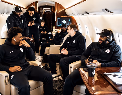 Six men in matching black athletic wear chatting and relaxing inside a private jet cabin.