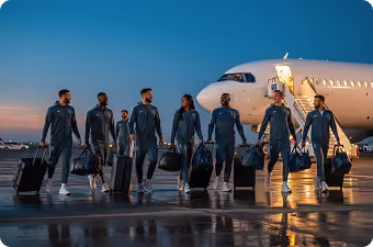 Group of athletes in matching tracksuits walking with luggage near a plane at dusk.