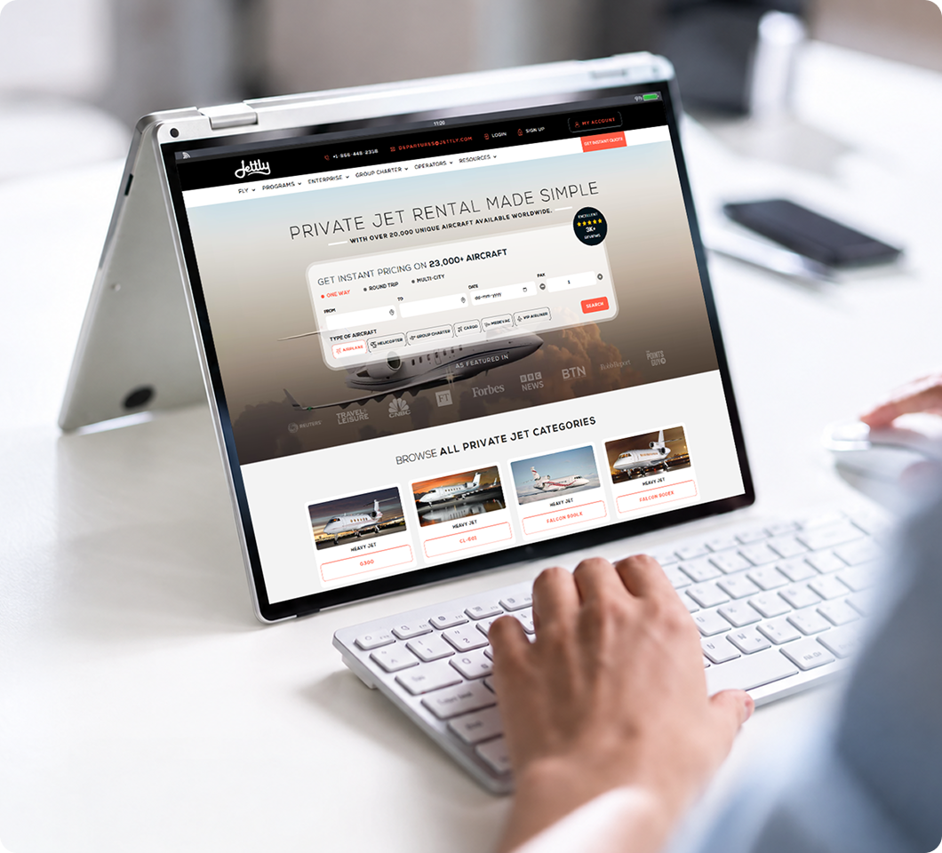 Person using a silver convertible laptop in tent mode on a white desk, browsing a private jet rental website showing options for one-way, round trip, and multi-city flights, types of aircraft, and categories of heavy jets including Gulfstream G300, CL-650, Falcon 900LX, and Falcon 900EX.