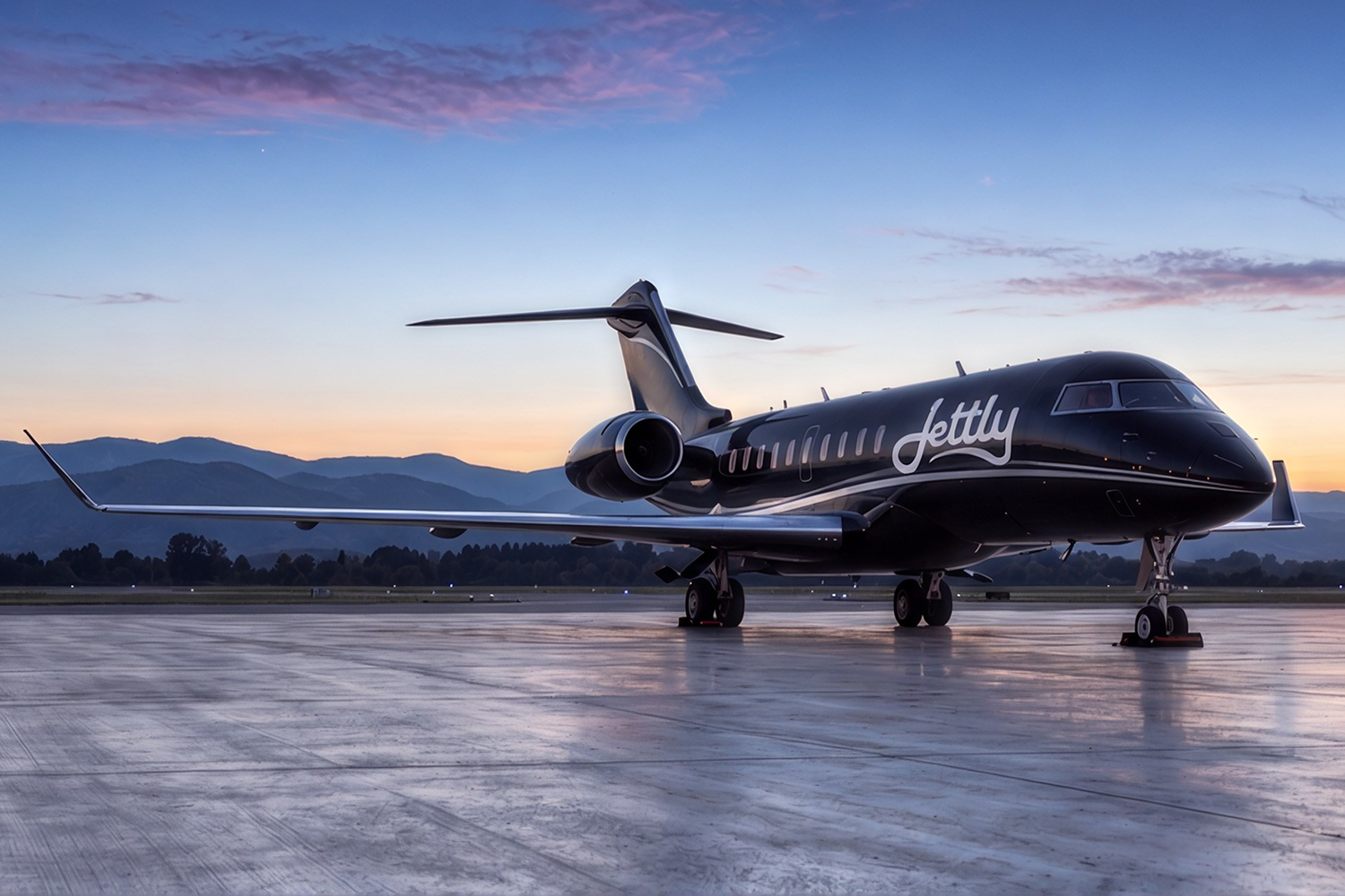 Sleek black private jet with the word 'Jettly' on its side parked on a wet tarmac at sunset with mountains and trees in the background under a partly cloudy sky.