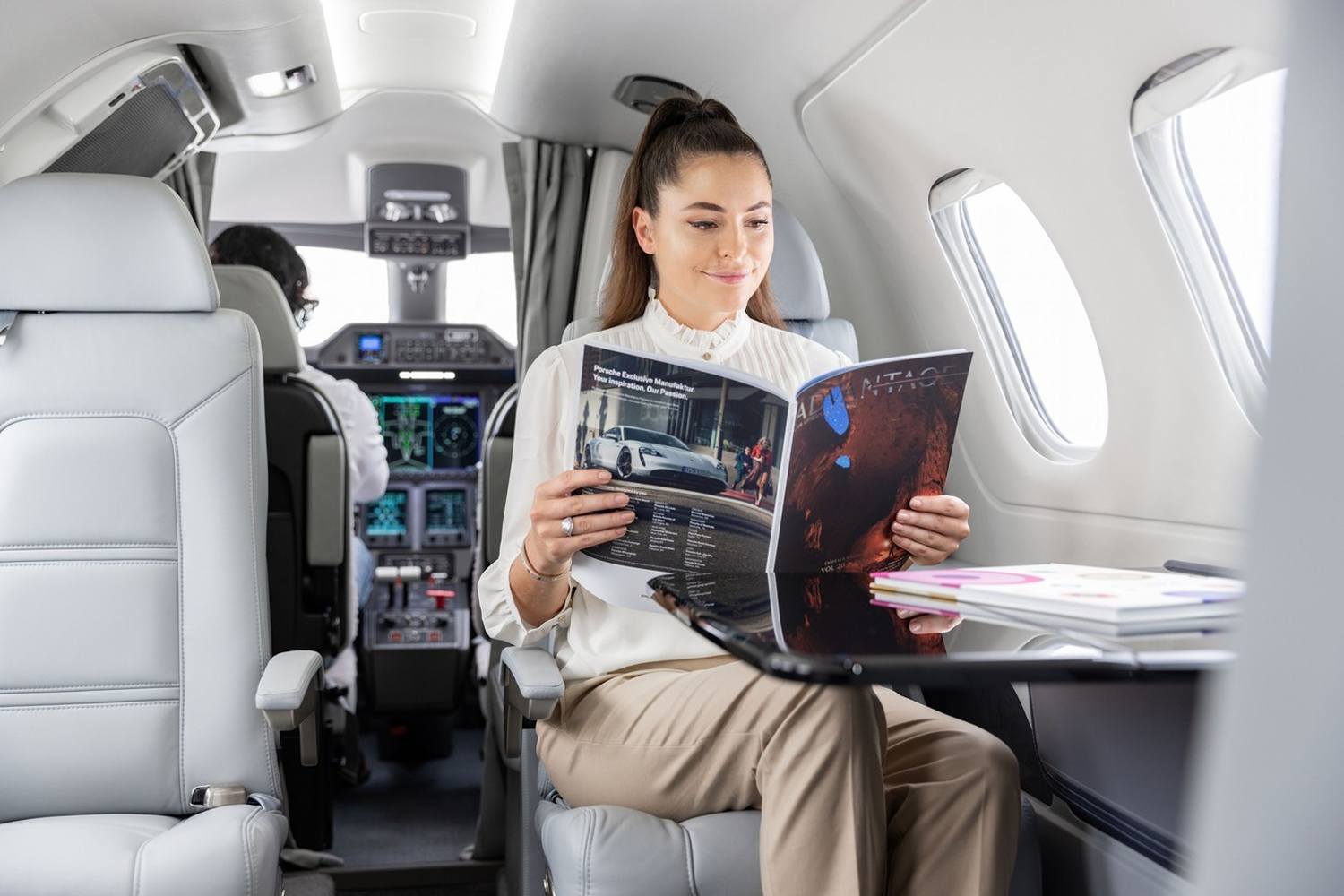 A woman with a ponytail wearing a white blouse and beige pants sits reading a magazine inside a private jet, with the cockpit and pilot visible in the background.