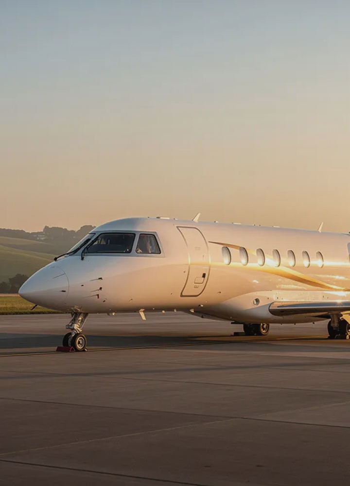 White private jet parked on runway at sunset with hills in the background, sunlight reflecting off its fuselage.