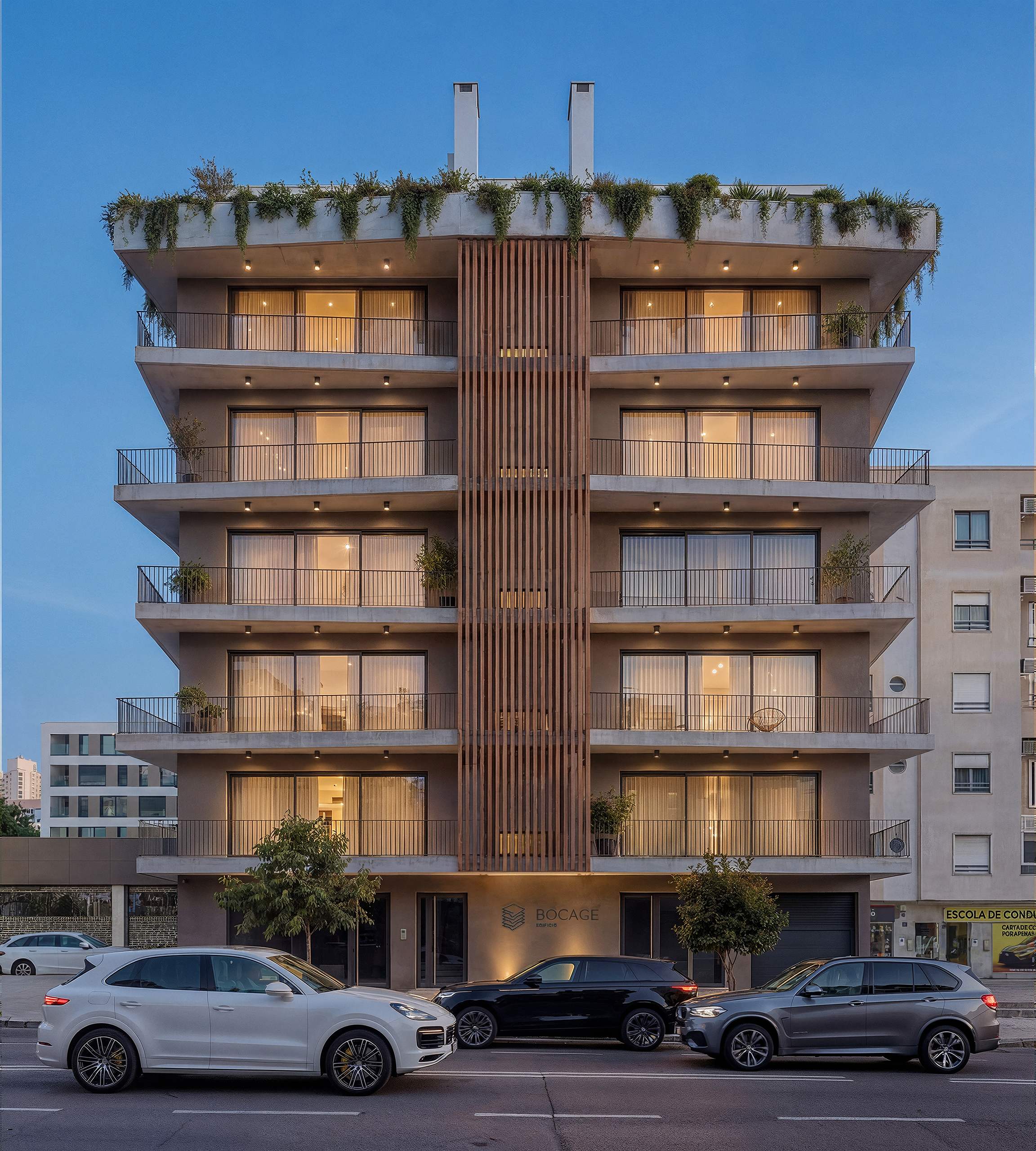 Fachada do Edifício Bocage ao anoitecer, com interiores iluminados e vegetação na cobertura.