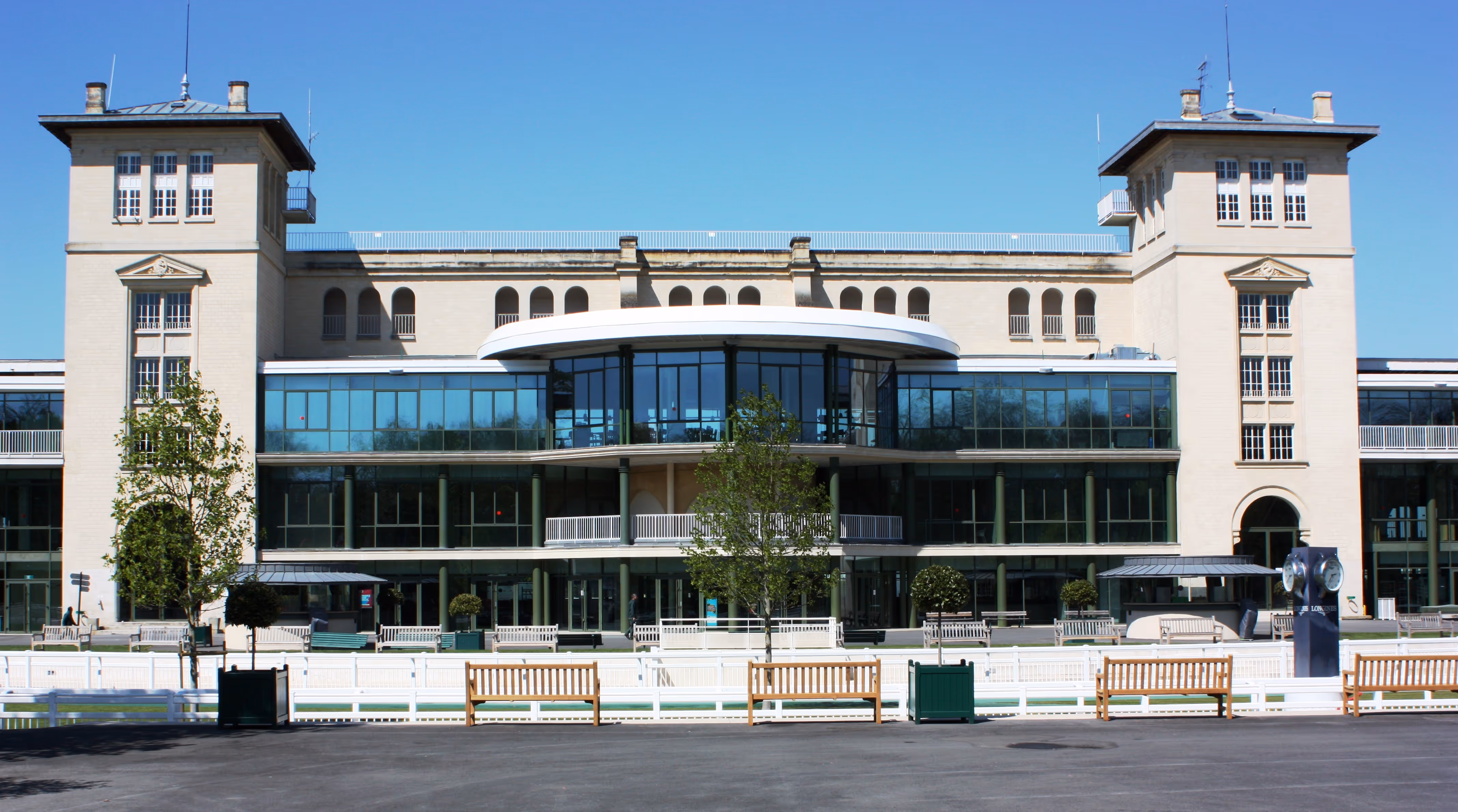 Large modern building with two tall towers, glass facade in the center, benches, and trees in front under a clear blue sky.