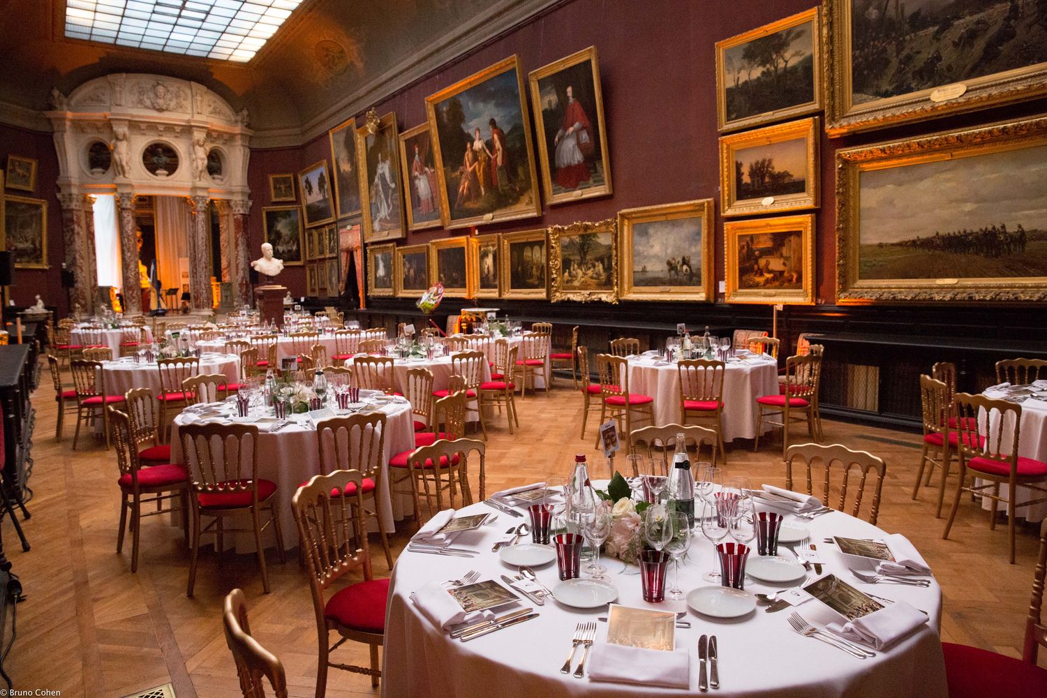 Elegant dining room with round tables set for a formal event, surrounded by classical paintings and a marble bust in a museum-like setting.