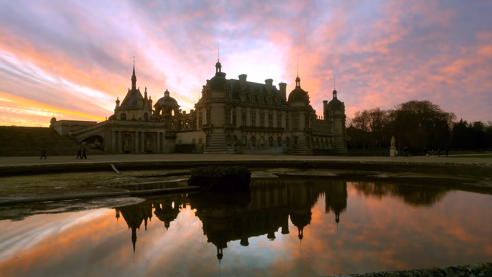 Historic chateau silhouetted against a colorful sunset sky with its reflection in a foreground pond.