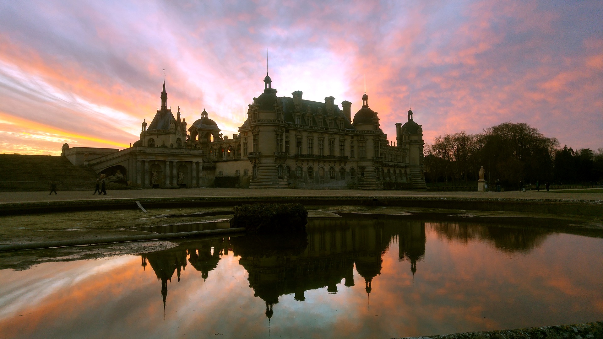 Historic chateau silhouetted against a colorful sunset sky with its reflection in a foreground pond.