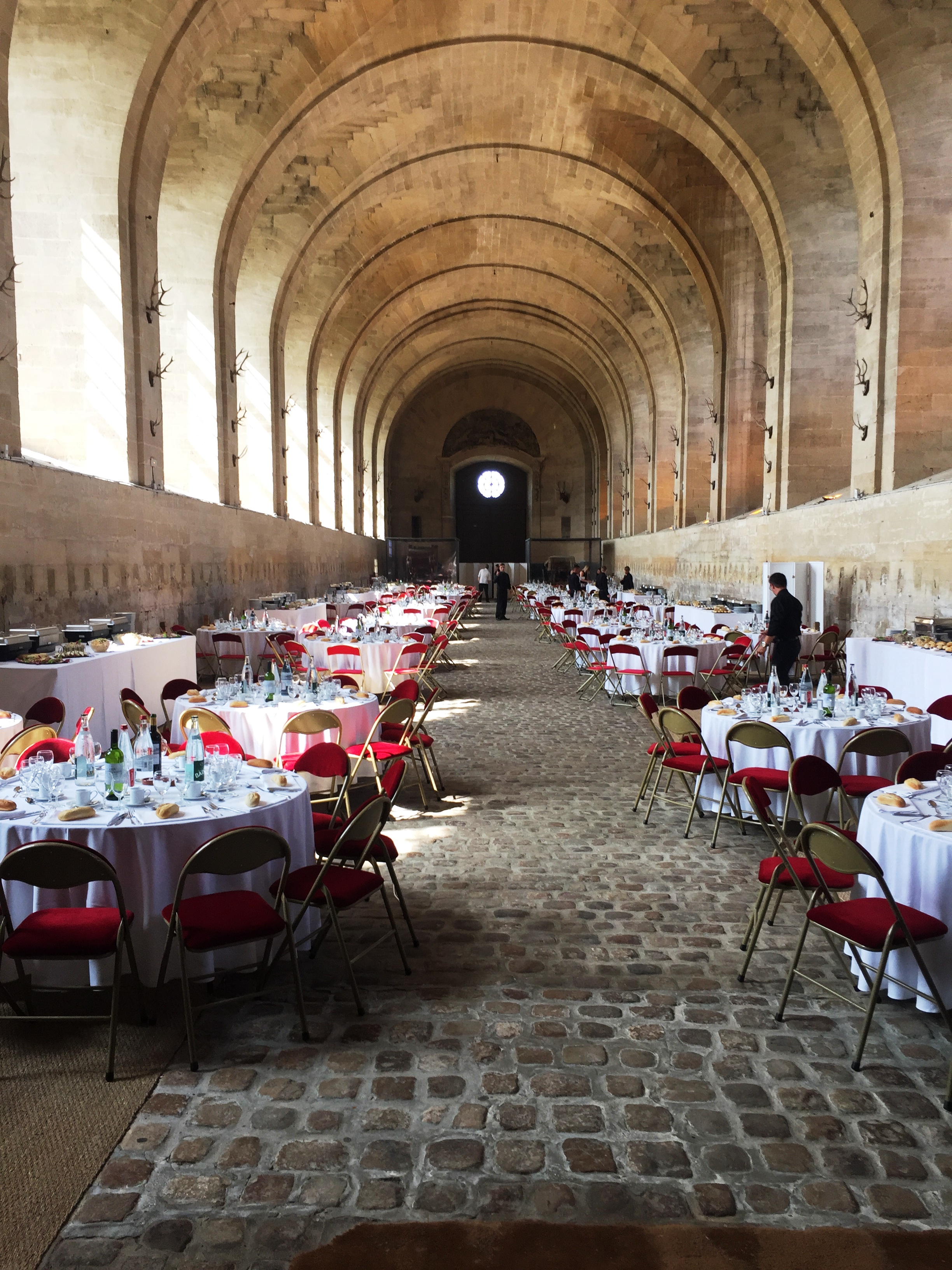 Long arched stone hall set up for a banquet with round tables covered in white cloth and red cushioned chairs.