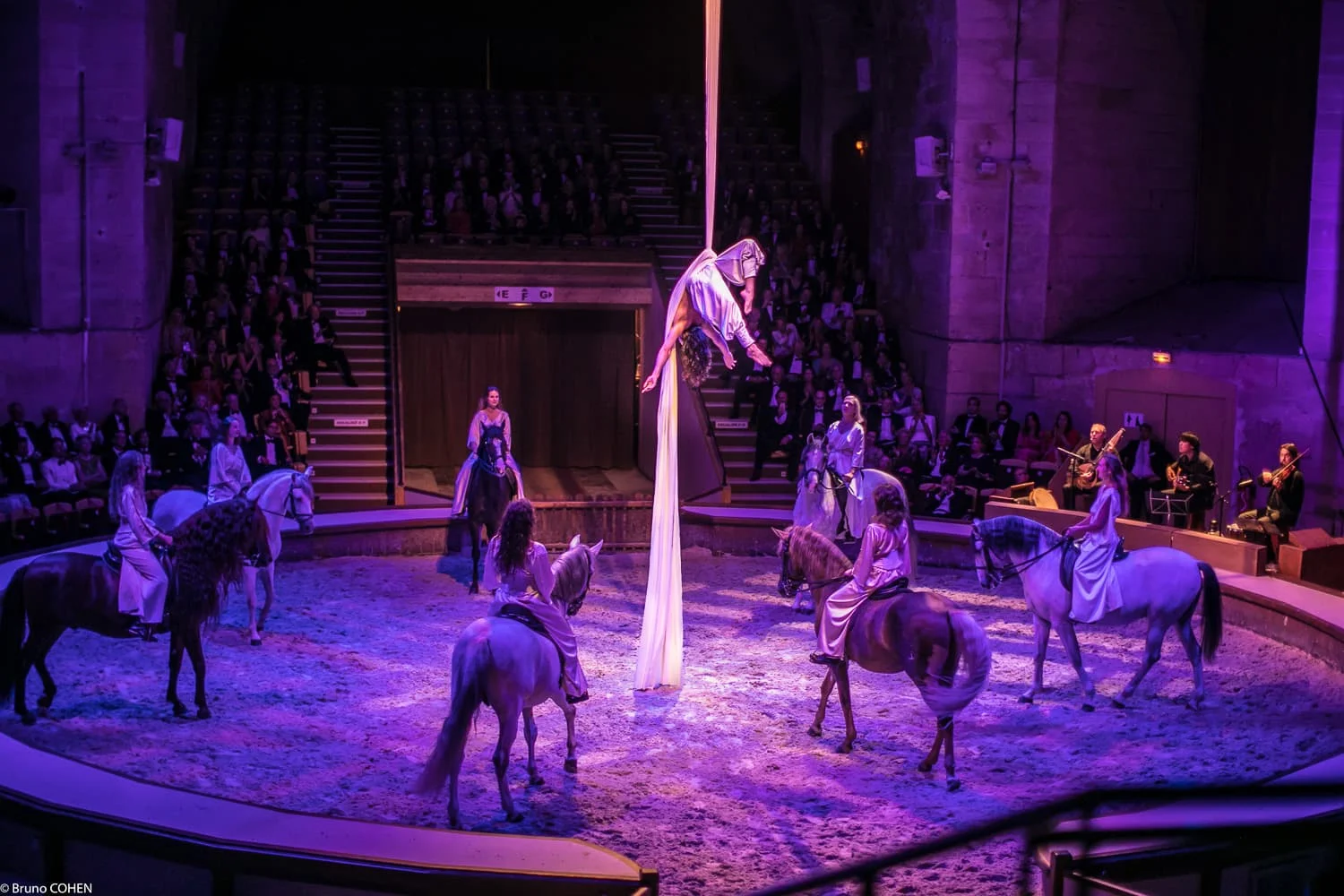 An indoor equestrian performance with six riders on horses surrounding an aerial silk artist performing in pink lighting.